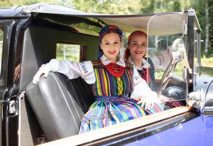 Dwie młode, uśmiechnięte kobiety  w kostiumach ludowych siedzą w środku zabytkowego samochodu. Zwrócone są do obiektywu