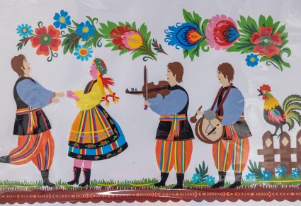 Prostokątna wycinanka łowicka ze scenką rodzajową, po lewej stronie tańczy para w kostiumie łowickim po prawej stoi dwóch chłopaków grają na instrumentach, obok nich na drewnianym płotku stoi kogut