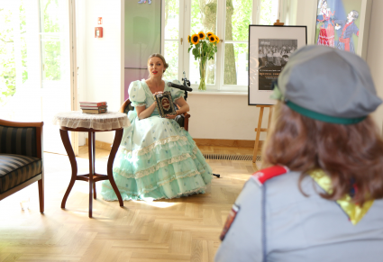 Przewodniczka Centrum Folkloru Polskiego "Karolin" siedząca przy stole w foyer Pałacu Karolin. W rękach trzyma lekturę "Nad Niemnem". Przed nią, patrząca w jej stronę, siedzi harcerka