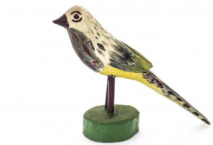 Drewniana figurka beżowego ptaka, ptak na brzuchu i ogonie ma bordowe pióra. Umieszczony jest na drewnianym patyku