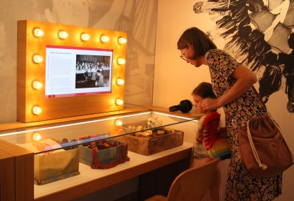 Sala ekspozycyjna, przy szklanej gablocie ze skrzynkami, w których są elementy kostiumów ludowych stoi dziewczynka i kobieta, przyglądają się gablocie. Na ścianie przed nimi znajduje się ekran z prezentacją