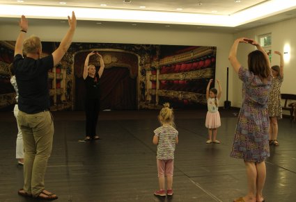 Sala prób, na parkiecie osoby dorosłe i dwie małe dziewczynki, ćwiczą taniec, mają uniesione ręce do góry, figura z baletu