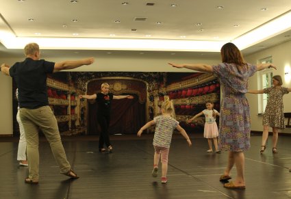 Sala prób, na parkiecie osoby dorosłe i małe dzieci, wszyscy uczą się układu tanecznego, powtarzają ruch za prowadzącą zajęcia