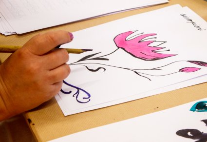 Zbliżenie na dłoń osoby, która trzyma w ręku bambusowy patyk do kaligrafii, przed nią leży kartka z namalowanym różowym kwiatem