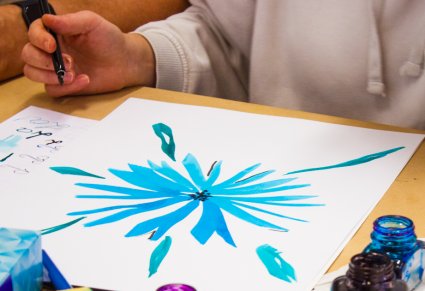 Zbliżenie na kartkę z namalowanym niebieskim kwiatem, na górze zdjęcia, po lewej stronie, dłoń kobiety, która trzyma pióro do kaligrafii