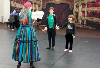Instruktorka tańca stoi po lewej stronie tyłem, przed nią stoi dziewczynka i chłopiec, trzymają się za ręce, wykonują ćwiczenie