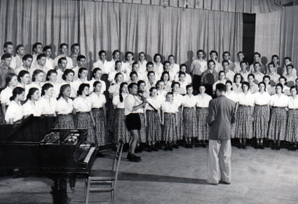 Sala prób, przed chórem Zespołu stoi Tadeusz Sygietyński, po lewej stronie na dole zdjęcia znajduje się fortepian