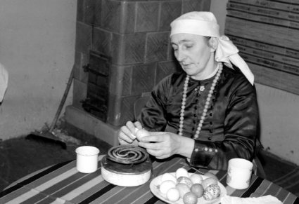 Kobieta siedzi przy stole, w ręku ma jajko, przed nią na stole jest talerz z gotowymi pisankami, stara kuchenka elektryczna i dwa kubki. Za nią w tle, piec kaflowy