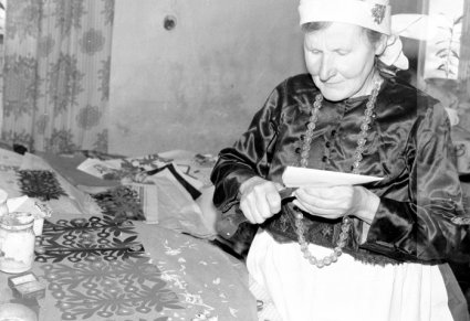 Starsza kobieta wycina nożyczkami z papieru, obok niej na stole leżą już gotowe wycinanki