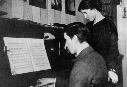 Przy fortepianie siedzi młody Tadeusz Sygietyński, obok niego stoi kobieta, na fortepianie nuty, na które wspólnie patrzą