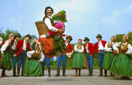A group of Mazowsze dancers watching a male dancer dressed up as an elderly lady. His head is sticking out of a wicker basket on her back
