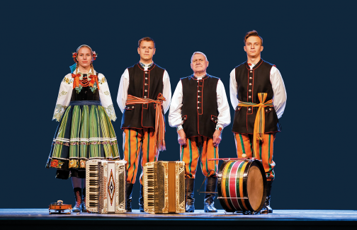 Four musicians standing on stage in costumes from the Łowicz region,  with their instruments placed on the floor in front of them
