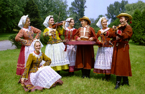 Siedmiu artystów zespołu Mazowsze w kostiumach rzeszowskich, w grupie pięciu dziewcząt jedna gra na skrzypcach, dwóch mężczyzn gra na cymbałach i skrzypcach