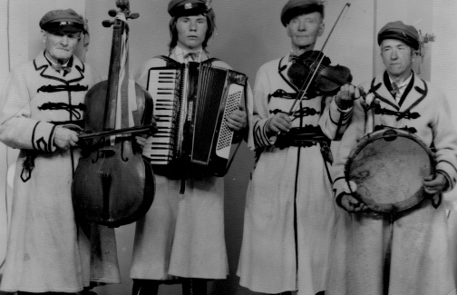 Czarno białe zdjęcie, na nim czterech mężczyzn trzymających instrumenty muzyczne. Mężczyźni ubrani są w kostiumy ludowe
