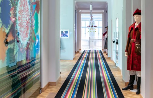 Korytarz w Centrum Folkloru Polskiego Karolin" na podłodze kolorowy pasiasty dywan, po lewej stoi manekin w stroju kosyniera, po prawej, na ścianie kolorowa grafika