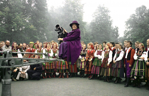Mira Zimińska-Sygietyńska na z kamerą siedzi na kranie filmowym, w tle stoją artyści zespołu Mazowsze