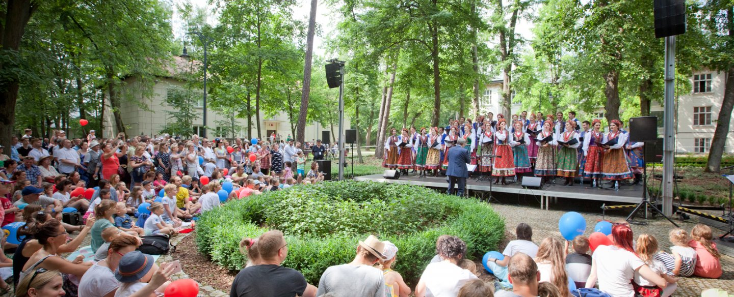 uczestnicy pikniku na siedzą na widowni przed zewnętrzną sceną w zielonym parku, na scenie chór zespołu Mazowsze
