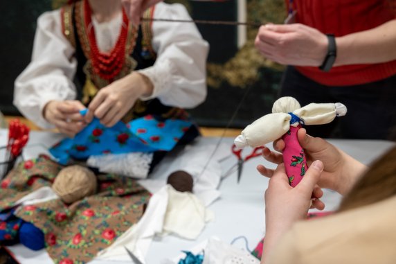 Zbliżenie na dłonie kobiet, trzymają skrawki materiałów formowane w lalkę, na stole warsztatowym leżą kolorowe materiały w kwiaty i nożyczki