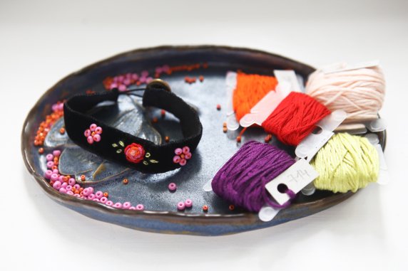 Na talerzyku leży bransoletka wykonana z aksamitu, koralików i nici, obok rozsypane koraliki po prawej na talerzyku leżą kolorowe nici