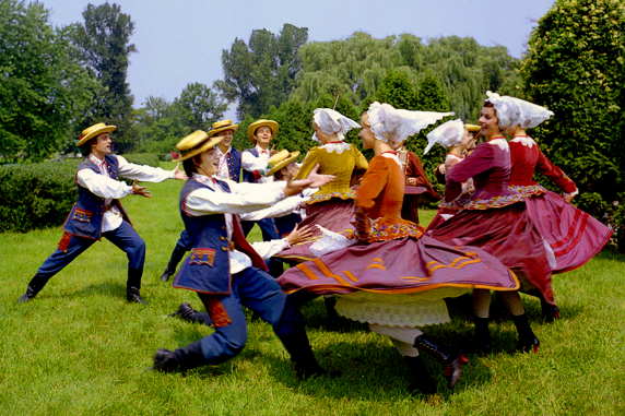 Grupa tańczących osób, pięć par w kostiumach rzeszowskich, za nimi muzycy z cymbałami i skrzypcami,  w tle park