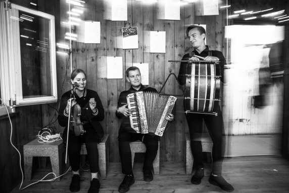 Three musicians sitting on stools in a small room. A young woman holds a violin one man is playing an accordion and one is carrying a drum