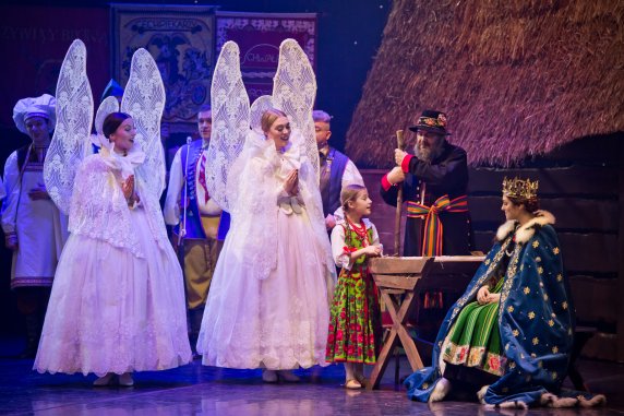 Spektakl Betlejem Polskie, na scenie dwie kobiety ubrane w kostiumy aniołów, mała dziewczynka w góralskim kostiumie oparta o kołyskę, nad kołyską Józef, obok siedzi Maria, oboje w kostiumie łowickim