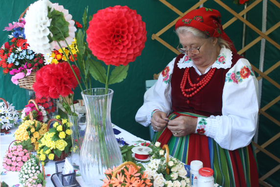 Przy stole siedzi starsza kobieta w kostiumie ludowym, skupiona trzyma dekorację w rękach. Na stole bukiety kolorowych kwiatów z bibuły
