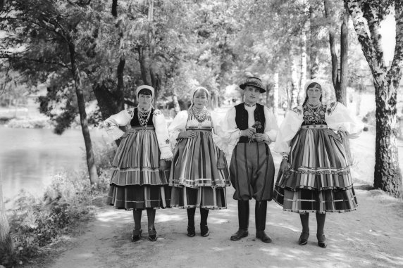 Czarno-białe zdjęcie, na nim trzy kobiety i jeden mężczyzna. Wszyscy ubrani w kostium łowicki. Stoją na tle parku, po lewej i prawej stronie za nimi rosną drzewa
