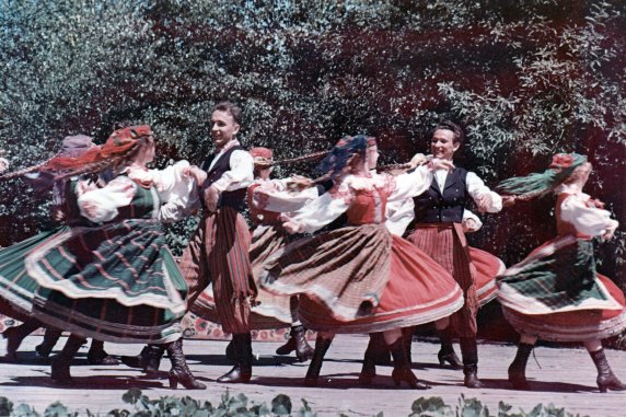 An archival photo of members of Mazowsze Ensemble in folk costume, dancing on the outdoor stage.