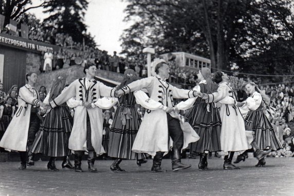 Czarno białe zdjęcie, na dworze grupa tancerzy z zespołu Mazowsze tańczy, za nimi na trybunach siedzi widownia