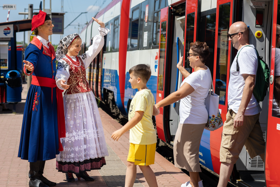 A family disembarking a Warsaw Commuter Railway train are greeted on the platform by Mazowsze artists wearing costumes from the Wilanow region