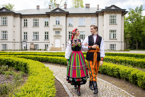 A couple dressed in folk costume from the Łowicz region walking through Karolin Park with the palace in the background