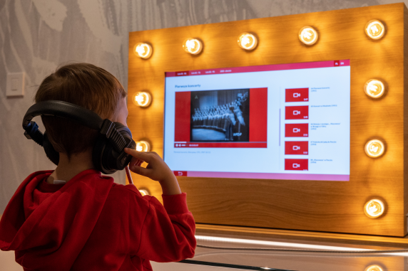Chłopiec na wystawie Centrum Folkloru Polskiego przy stanowisku multimedialnym zakładający słuchawki