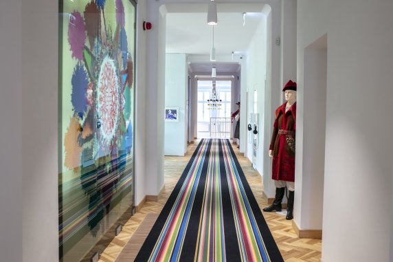 Korytarz we wnętrzach Pałacu Karolin, na podłodze kolorowy, pasiasty dywan, po lewej stronie manekin ubrany w kostium kosyniera.