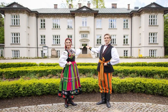 A smiling couple in costumes from the Łowicz region standing in front of Karolin Palace gesture towards the open front door, inviting visitors into the Centre for Polish Folklore