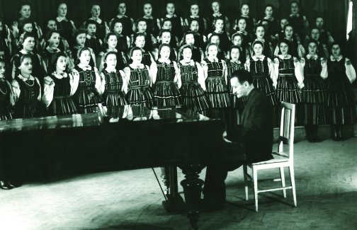 Tadeusz Sygietyński gra na fortepianie, z tyłu stoi chór zespołu "Mazowsze" ubrany w piękne kostiumy, zdjęcie archiwalne czarno-białe.