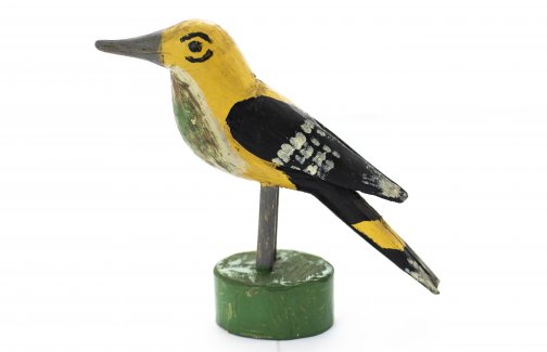 Drewniana figurka żółtego ptaka, ptak na brzuchu i ogonie ma czarne pióra. Umieszczony jest na drewnianym patyku.