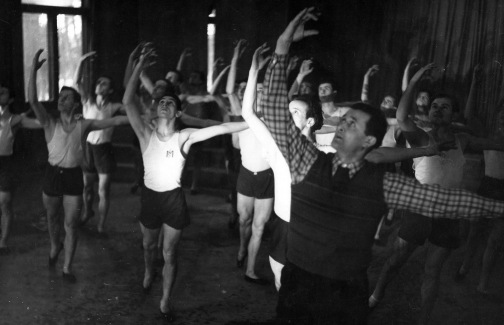 Czarno białe zdjęcie, grupa mężczyzn ćwiczy balet. Mają uniesione prawe dłonie do góry, wzrok uniesiony za dłonią